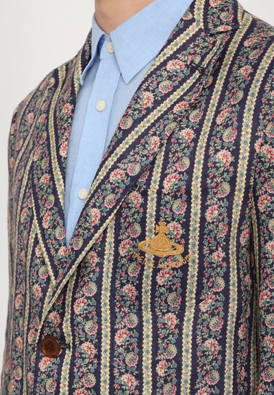 Floral patterned blazer with navy and cream stripes, featuring a brown button and gold embroidered emblem. Worn over a light blue shirt.