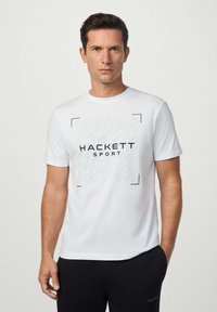 Bílá tričko s krátkým rukávem a čern�ým logem "HACKETT SPORT" ve středu, s geometrickým vzorem na pozadí. Měkká tkanina.