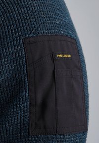 Der gestrickte, dunkelblaue Pullover hat ein strukturiertes, gewebtes Material mit einer kontrastierenden schwarzen Stofftasche, die mit "PME LEGEND" in Gelb beschriftet ist.