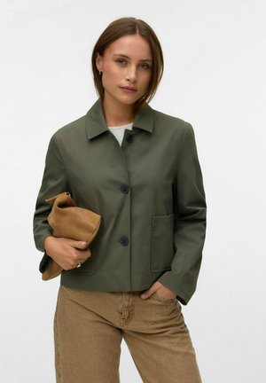 Femme portant une veste boutonnée vert olive et un pantalon marron tient une pochette beige pliée dans une main sur un fond blanc uni.