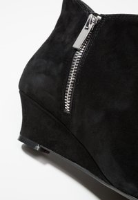 Botte de chevilles en suede noir avec un talon compensé, dotée d'une fermeture éclair argentée sur le côté et d'une surface texturée lisse.