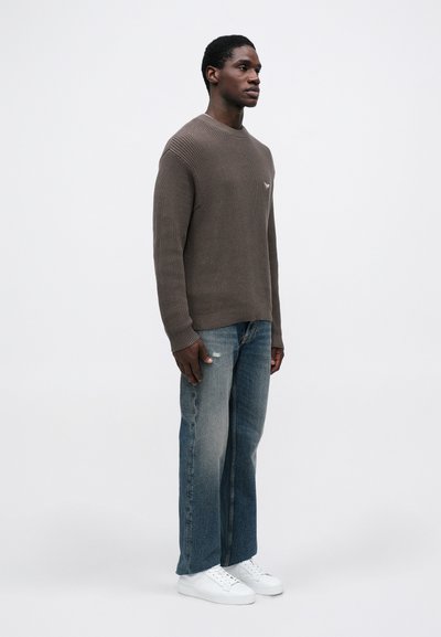 Rjav rebrast pulover z okroglim izrezom, v kombinaciji z uničenimi modrimi džinsom in belimi športnimi copati, s sproščenim krojem in minimalnim dizajnom.