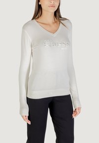 Lichtgrijze V-halstrui van zacht materiaal, met een geborduurd "Guess" logo in zilver, en geribbelde manchetten en zoom. Gemaakt in combinatie met zwarte broek.