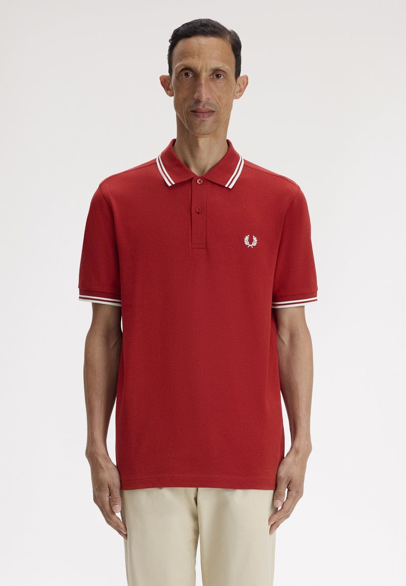 Uomo in piedi, rivolto in avanti, che indossa una polo rossa con strisce bianche sul colletto e sulle maniche, e pantaloni beige.