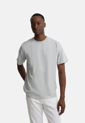Hombre de piel oscura que lleva una camiseta de manga corta gris claro y pantalones blancos, de pie con una mano detrás de la espalda sobre un fondo blanco.
