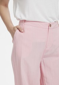 Růžové lněné kalhoty s rovnýma nohama, zapínáním na knoflík vpředu a postranními kapsami. Látka má jemný, mírně texturovaný vzhled.