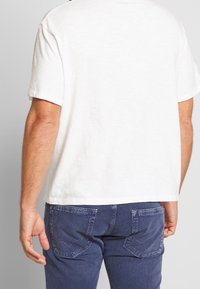 Bílý bavlněný tričko s krátkými rukávy, zobrazené zezadu. V kombinaci s modrými džíny se šitými detaily na kapsách. Pohodlný střih.