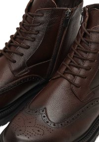 Bottes en cuir marron avec un design à lacets et une fermeture éclair sur le côté. Détails de brogue remarquables le long des bords et surface texturée.