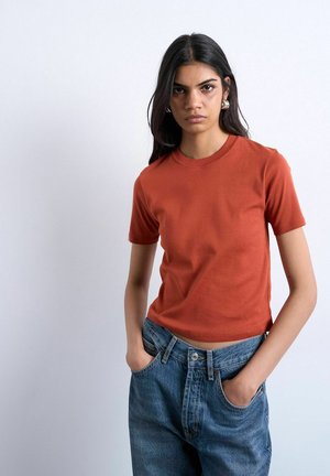 Young woman with long dark hair wearing a rust-colored short-sleeve shirt and blue jeans, standing with hands in pockets against a plain background.