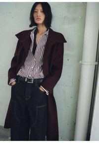 Manteau en laine bordeaux avec un grand col, chemise rayée rouge et blanche en dessous, jeans larges noirs avec des coutures jaunes, et ceinture noire.