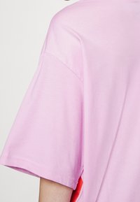 Camiseta de algodón rosa con un corte holgado, que presenta mangas cortas y aberturas laterales que revelan un interior rojo en contraste. Textura suave.