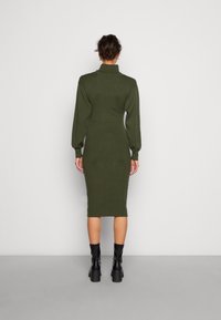 Robe en maille vert olive avec un col montant et des manches bouffantes, coupe ajustée, longueur genou, associée à des bottes à talons noires.