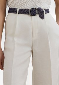 Woven navy belt with a metal square buckle worn over cream trousers; textured fabric with a braided pattern visible across the belt.