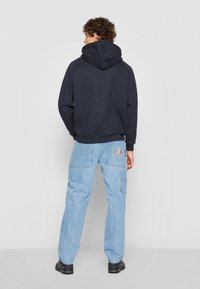 Felpa con cappuccio blu navy e tasca a marsupio, abbinata a pantaloni cargo in denim azzurro chiaro con un dettaglio ricamato a forma di arcobaleno. Scarpe nere.