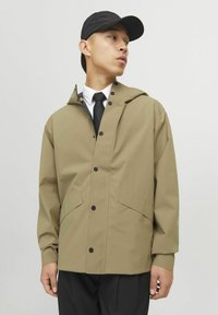 Beige waterdichte jas met een capuchon, drukknoopsluitingen en twee voorzakken. Gestyled met een zwarte pet en een wit shirt, met een zwarte stropdas.