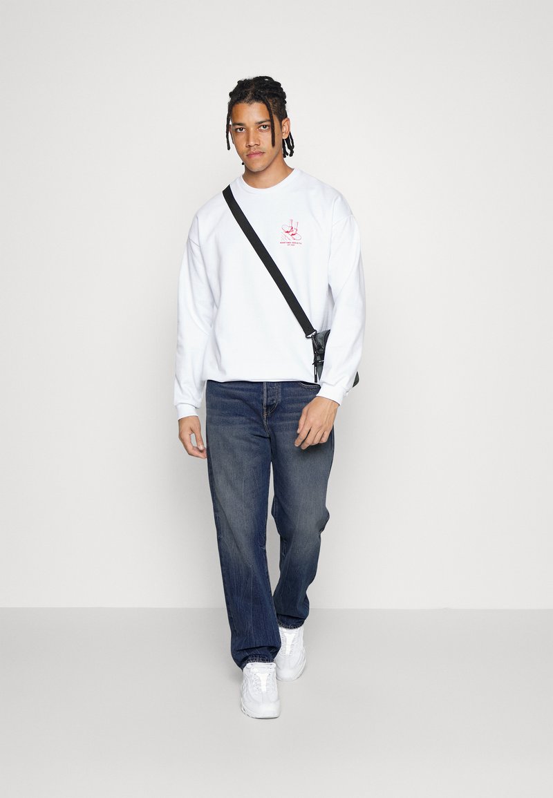 Weißes Sweatshirt mit rotem Grafikdesign, kombiniert mit dunkelblauen Denim-Jeans und weißen Sneakers. Das Modell trägt eine schwarze Umhängetasche.