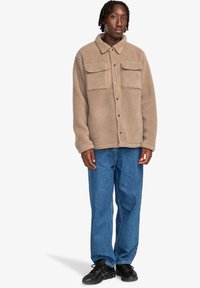 Beige fleece jacket with two chest pockets, button closure, and a collar, paired with blue denim pants and black sneakers.