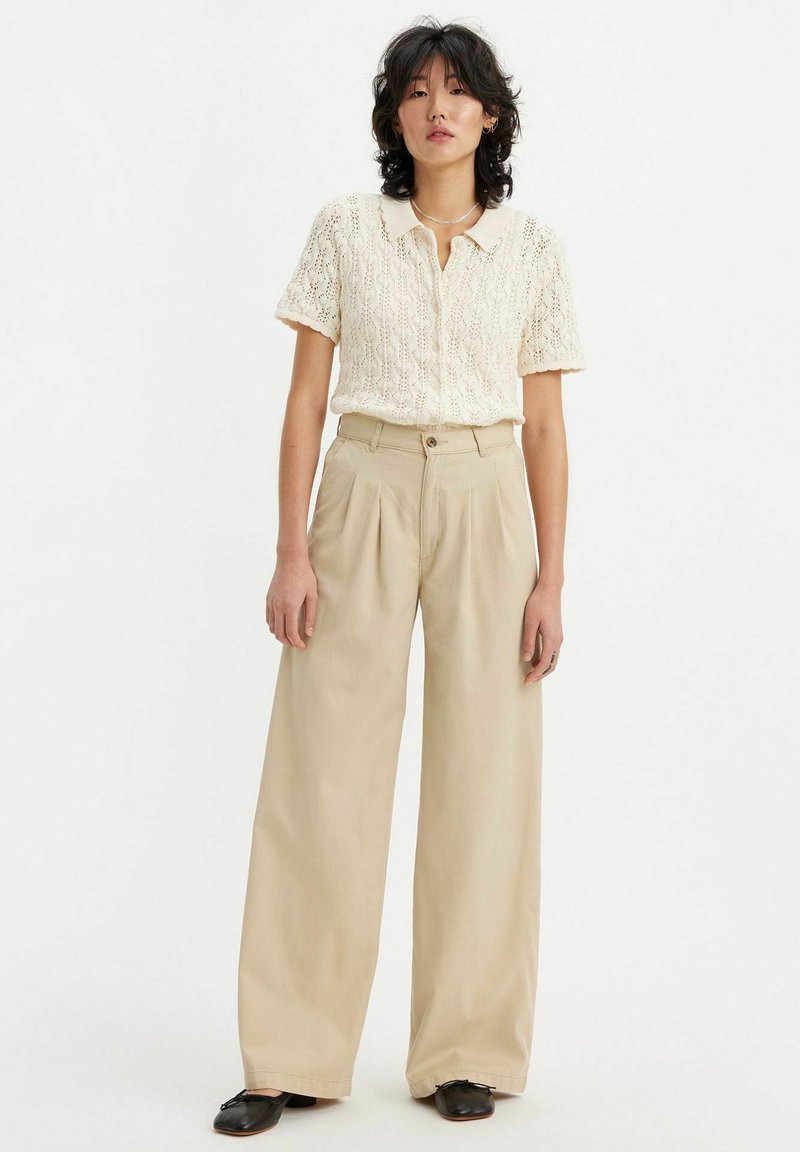 Beige hoge taille broek met wijde pijpen en plooien, gecombineerd met een crèmekleurige, gestructureerde, korte mouwen gebreide cardigan. Zwarte schoenen.