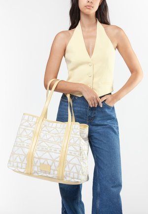 Mujer con chaleco halter amarillo pálido y vaqueros azules sosteniendo un gran bolso tote blanco y beige con asas largas.
