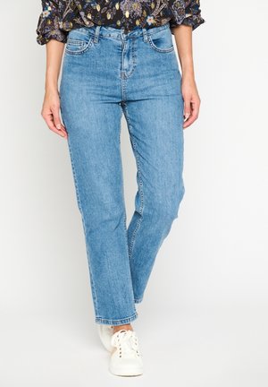 Femme portant un jean droit bleu clair, un chemisier à manches longues à fleurs rentré, et des baskets blanches, debout sur un fond uni.