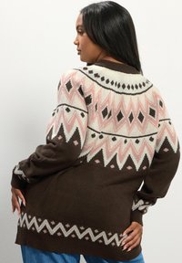 Suéter de punto marrón con diseños y patrones geométricos en rosa y crema. Presenta un cuello y puños acanalados, mangas largas y un ajuste relajado.