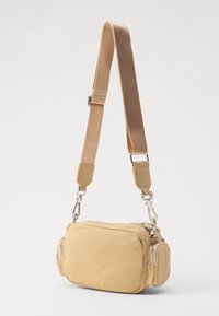 Borsa a tracolla beige di forma rettangolare, realizzata in tessuto morbido. Presenta una chiusura a zip, due tasche laterali e una tracolla regolabile.