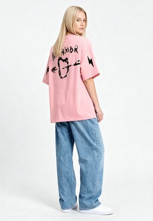 Jeune femme aux longs cheveux blonds portant un t-shirt rose ample avec des graphismes noirs et un jean large bleu clair, debout et regardant derrière elle.