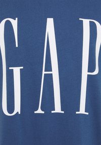 T-shirt en coton bleu avec un grand texte blanc épelé "GAP". Le tissu a une texture lisse avec une coupe décontractée et des éléments de design minimalistes.