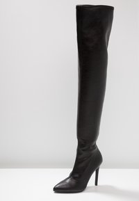 Botte noire au-dessus du genou en cuir lisse, avec un bout pointu et un talon aigu haut. Des coutures décoratives traversent l'avant.