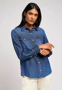 Camicia in denim blu scuro con chiusura a bottoni sul davanti, colletto a punta, tasche sul petto e maniche lunghe, con bottoni a pressione bianchi e orlo curvato.