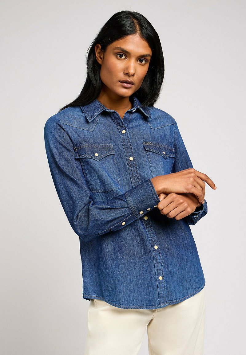 Camicia in denim blu scuro con chiusura a bottoni sul davanti, colletto a punta, tasche sul petto e maniche lunghe, con bottoni a pressione bianchi e orlo curvato.