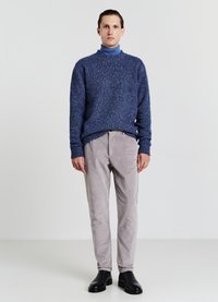 Maglione lavorato a maglia blu con collo alto, abbinato a pantaloni in velluto a coste grigi chiari e stivaletti neri. Modello in posizione eretta su uno sfondo neutro.