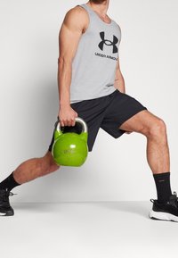 Canottiera grigia senza maniche con logo nero, pantaloni corti sportivi neri, tiene un kettlebell verde, sneakers nere, in posizione di affondo.