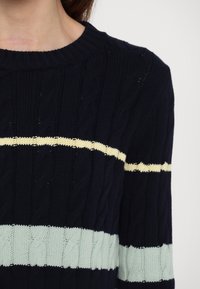 Personne portant un pull bleu marine en maille torsadée avec des rayures horizontales crème et bleu clair sur la poitrine et les manches.