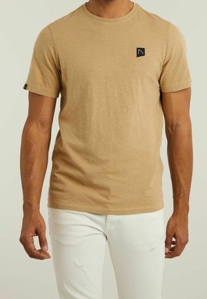 Maglietta a maniche corte color tan realizzata in tessuto morbido, con un piccolo logo nero ricamato sul petto, abbinata a jeans bianchi strappati.