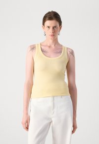Yellow ribbed tank top with a rounded neckline, fitted design, and subtle logo detail, paired with white trousers. Smooth texture.