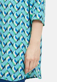Turquoise blouse met een geometrisch patroon met blauwe en groene vormen. Korte mouwen en een losse pasvorm, gemaakt van lichte stof.