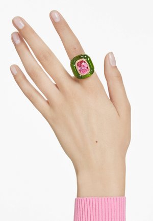 A hand wearing a bold green ring featuring a large pink gemstone. The hand rests against a light background, with a pink knit cuff visible.