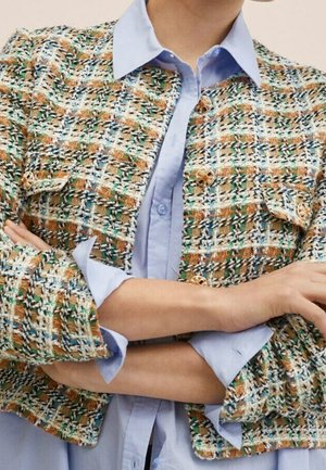 Personne portant une chemise bleue à boutons sous une veste en tweed multicolore avec des boutons dorés, les bras croisés sur la poitrine.