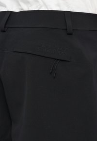 Fekete nadrág sima textúrával, húzózsinórral és hátul egy kis hímzett logóval, amely a nadrág színével megegyező szálat használ.