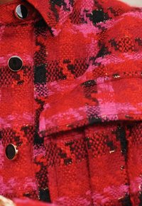 Punainen ja vaaleanpunainen kuvioitu tekstiilitakki, jossa on tunnusteltava rakenne, mustat napit ja etutaskun yksityiskohta.