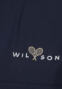 Un textil azul marino presenta la palabra "WILSON" en bordado blanco, con dos raquetas de tenis cruzadas en hilo dorado debajo.