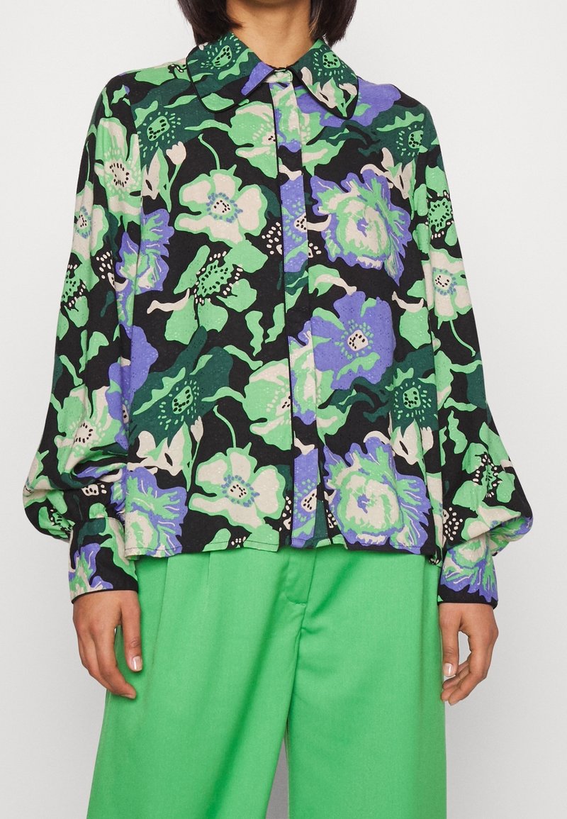 Femme portant un chemisier à manches longues à motif floral avec des couleurs vert, violet et noir, associé à un pantalon vert vif.
