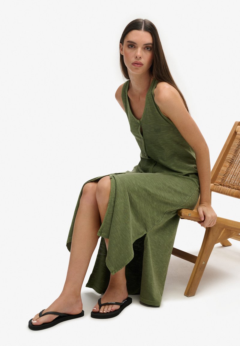 Robe maxi verte sans manches avec un devant boutonné, une silhouette fluide et une fente latérale haute ; associée à des tongs noires sur une chaise en bois.