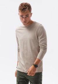 Giovane uomo con capelli ricci, che indossa una camicia a maniche lunghe beige, pantaloni verdi e un orologio da polso nero, che guarda dritto davanti a sé su uno sfondo bianco.