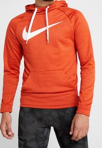 Sweat à capuche orange en tissu texturé, avec un grand logo Nike blanc sur la poitrine et des cordons blancs. Comprend une poche avant.