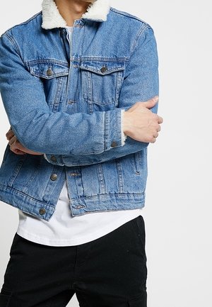 Homme portant une veste en denim bleu avec col en polaire blanche, chemise blanche et pantalon noir, les bras croisés sur la poitrine.