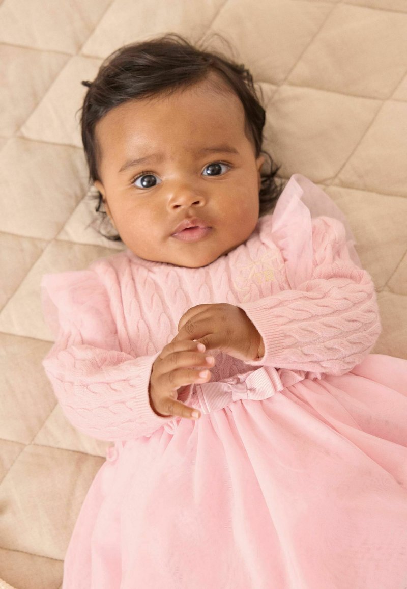 Vauva, jolla on tummat hiukset ja ruskea iho, makaa beige-värisellä torkkupeitteellä, päällään vaaleanpunainen mekko, jossa on neulotut hihat ja vyötäröllä rusetti, katse suunnattuna eteenpäin.