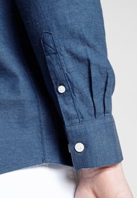 Manche de chemise en denim bleu avec deux boutons blancs sur le poignet et des plis, portée sur un poignet au-dessus de vêtements blancs.