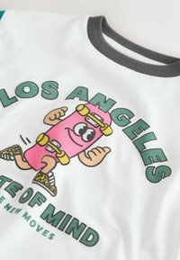 Valkoinen t-paita, jossa on sarjakuvatyylinen pinkki skeittihahmo keltaisilla yksityiskohdilla ja ilmeikkäillä silmillä, jota ympäröi vihreä teksti. Tumma, ribbineulottu kaulus.